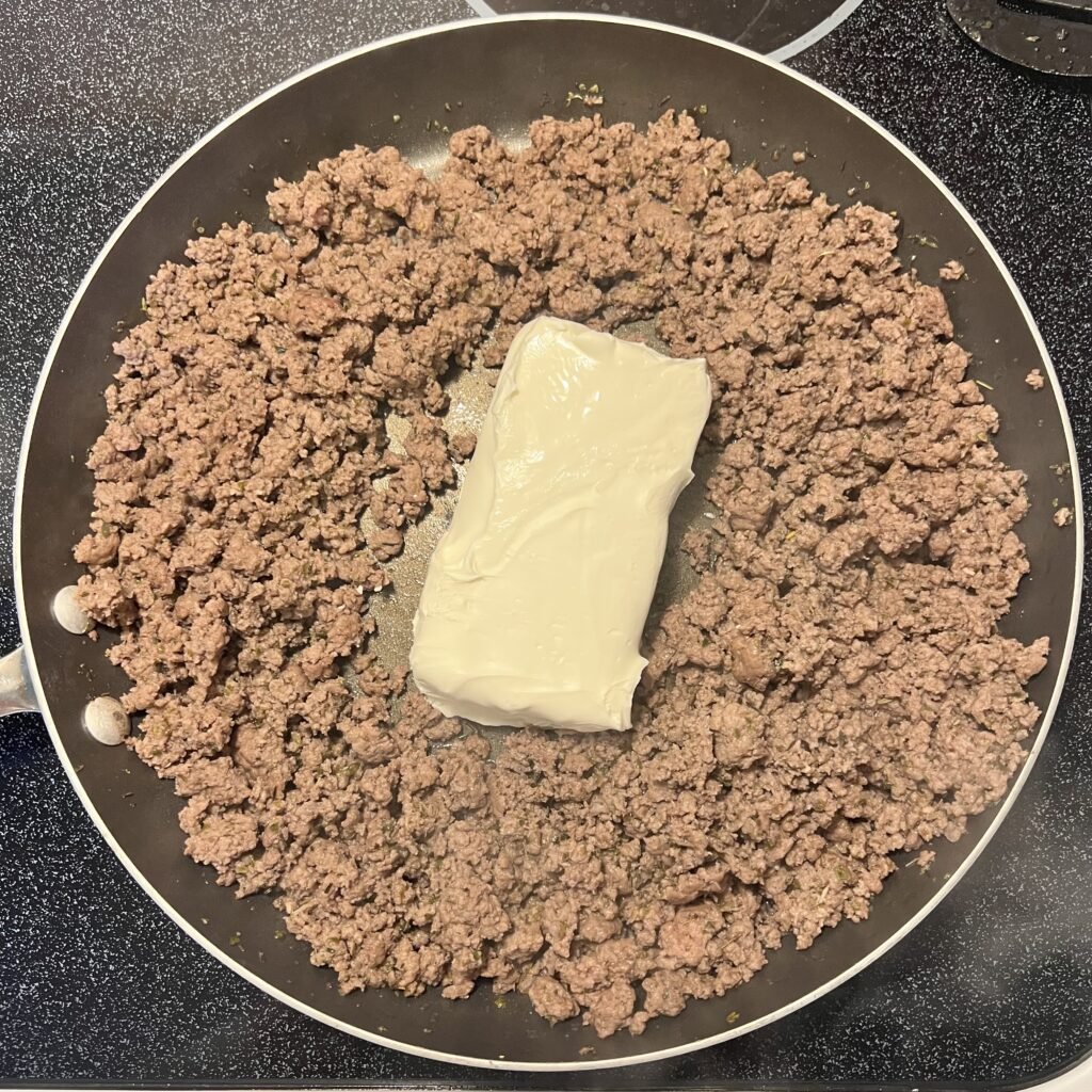 A block of cream cheese in the center of a frying pan surrounded by browned ground beef. This is the second step to making creamy ground beef.