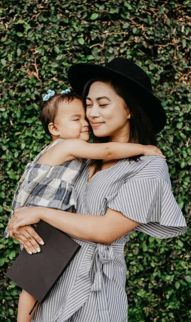 A mom and her daughter hugging and smiling. They are standing in front of beautiful greenery with light shining up on them.