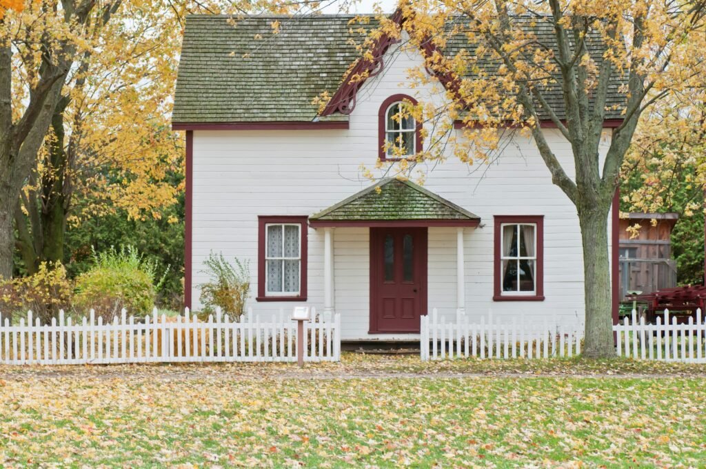 The front of a beautiful home with white siding and burgundy trim sitting peacefully amongst many trees in fall, with their rich yellow and orange leaves.