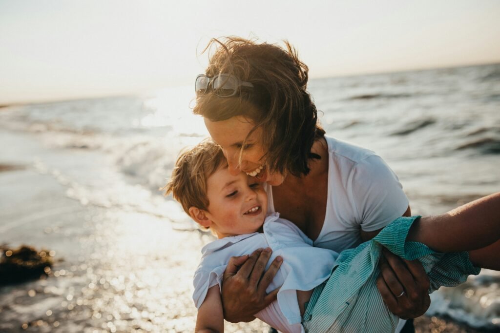 A mom holding her son, laughing and playing by the ocean. The sun is shining in the background.