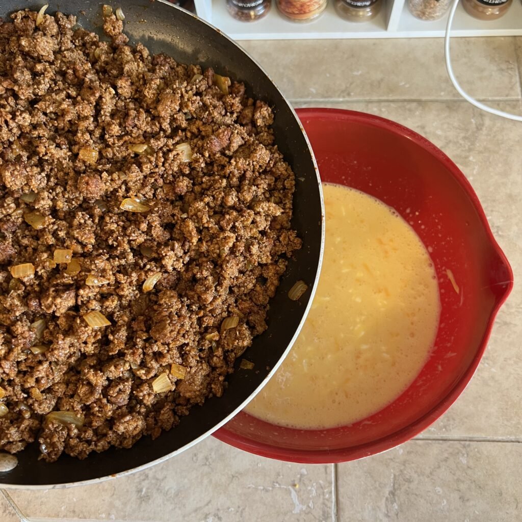 Taco and onion ground beef over a bowl of an egg mixture about to be mixed together to make a dinner casserole.