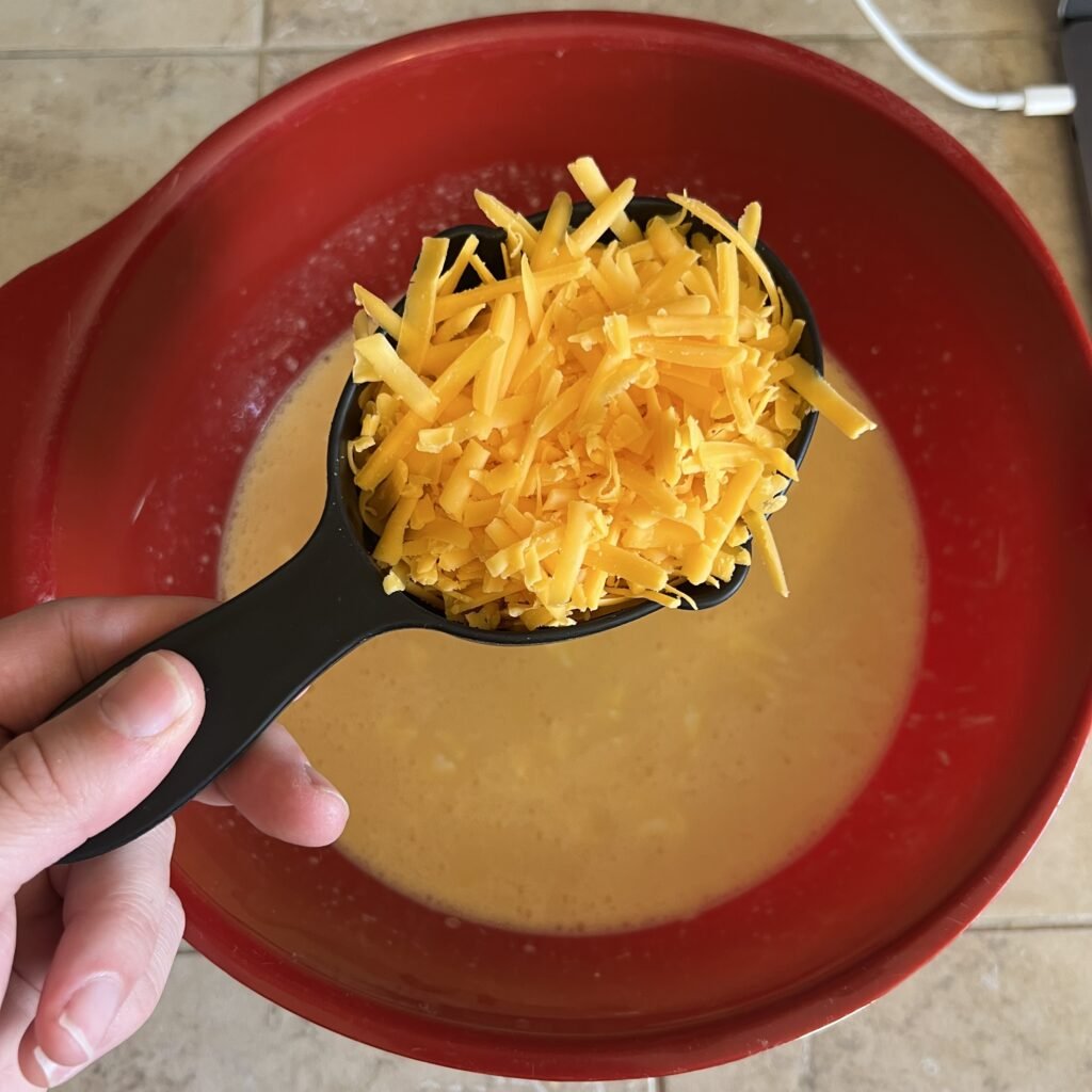 An overhead shot of a cup of shredded cheddar cheese over a bowl of eggs mixture for a simple dinner casserole.