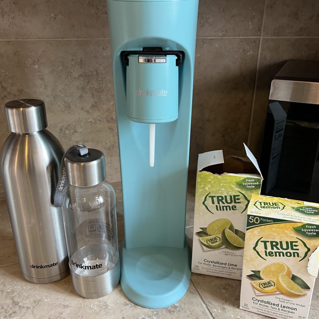 A turquoise blue Drinkmate pictured with two Drinkmate bottles, a box of True Lime, and a box of True Lemon.