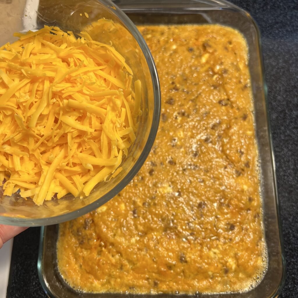 An overhead shot of a bowl of shredded cheese with a baked casserole in the background.