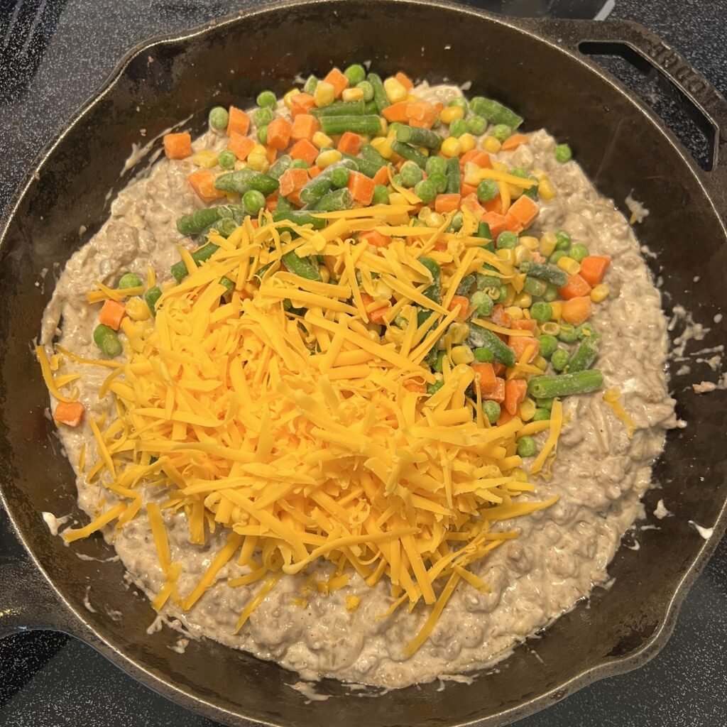 Adding the veggies and shredded cheddar cheese to the casserole filling.