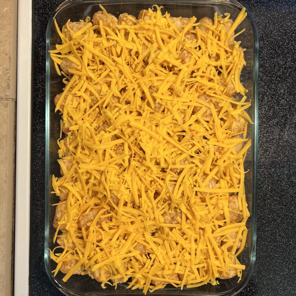 Shredded cheddar cheese evenly sprinkled across the top of a tater tot hot dish, ready to bake to perfection in the oven.