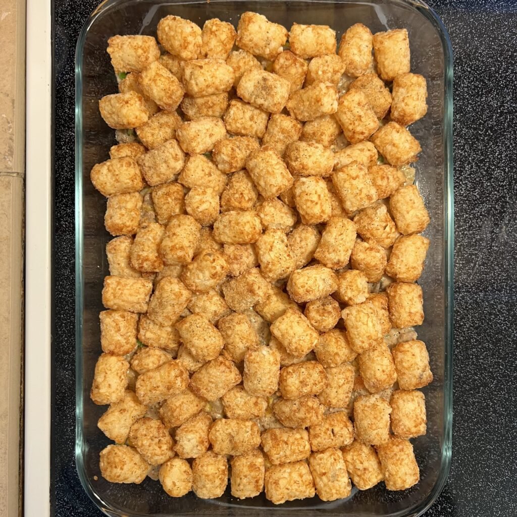 Tater tots spread out over the casserole filling, the second step in assembling this yummy hot dish.